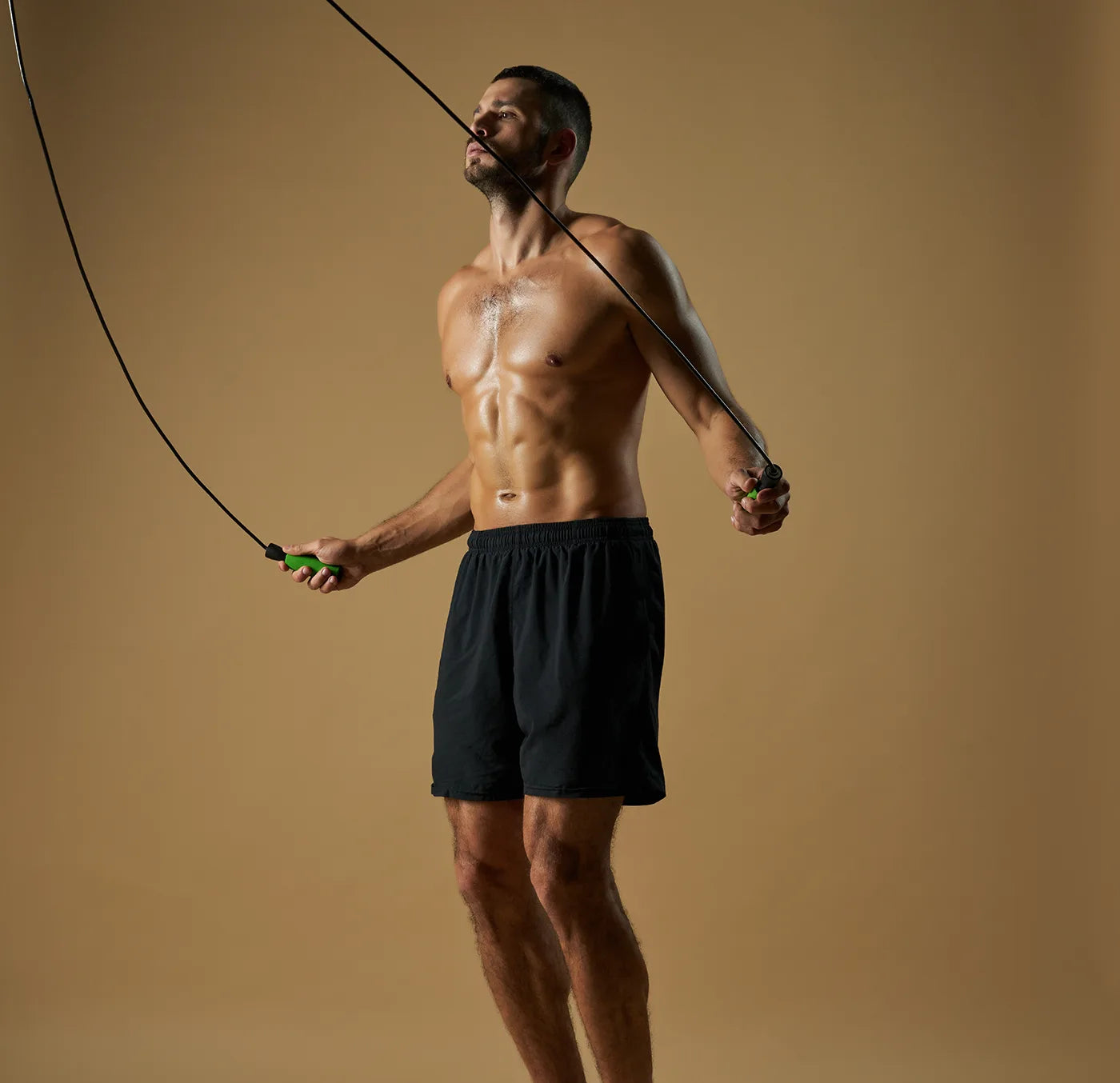 A shirtless man in black shorts is jumping rope against a plain light brown background. He appears focused and athletic, with light sweat visible on his upper body.