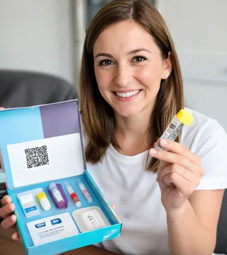A smiling woman in a white shirt holds up a vial and displays an open at-home medical test kit containing various tubes, containers, and a card with a QR code.