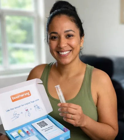 A woman with dark hair in a bun, wearing a green tank top, smiles while holding a test tube. In front of her is an open boomerang-branded at-home test kit with instructions and test tubes visible.