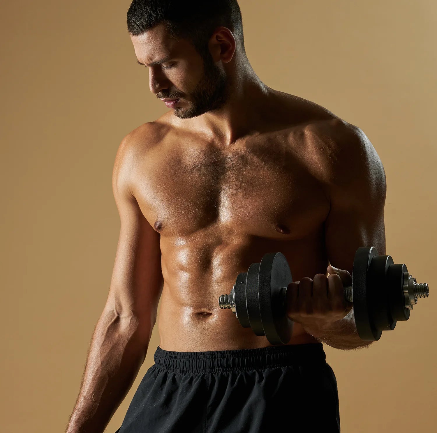 A muscular, shirtless man with short hair is lifting a dumbbell with his right arm. He is looking down at his arm and wearing black athletic shorts. The background is plain and beige.