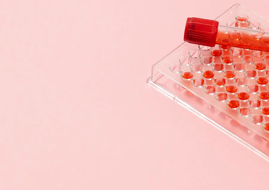A test tube filled with red liquid rests on a clear plastic microplate containing multiple wells, each holding a small amount of red liquid, against a pink background.