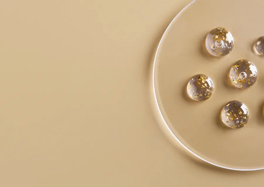 Clear gel spheres with gold flakes rest on a transparent round plate, placed on a beige surface with minimalist and elegant composition.