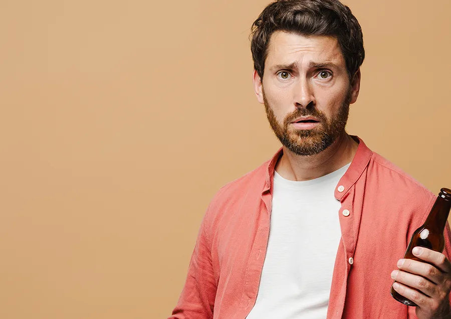 A man with short brown hair and a beard, wearing a white t-shirt and an open red shirt, holds a brown bottle in one hand and looks surprised or confused against a plain beige background.