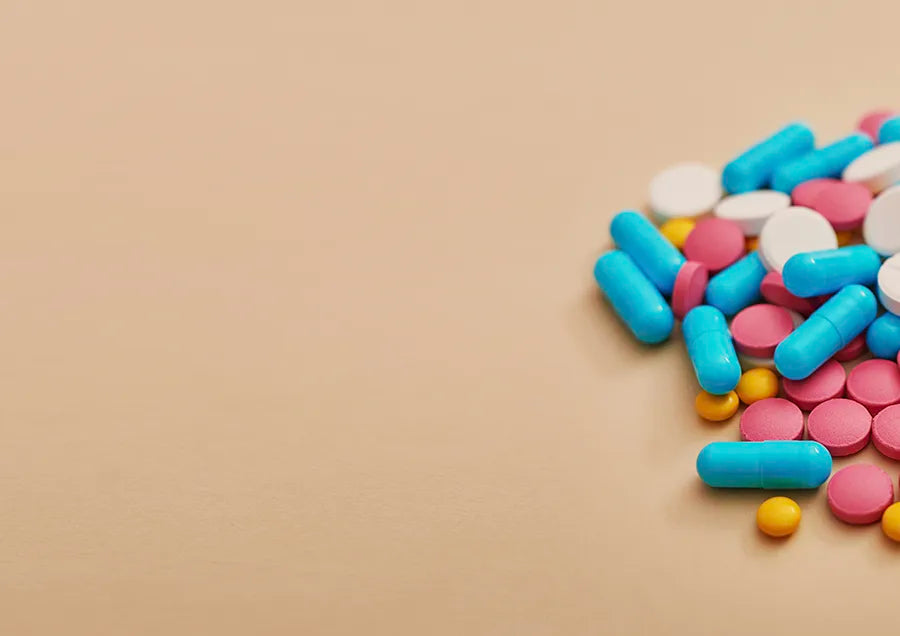 A variety of pills and capsules, including blue, pink, white, and yellow tablets, are scattered on the right side of a beige surface, with empty space on the left.