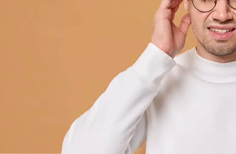 A man wearing a white sweater and glasses touches his head, partially smiling, with a beige background. Only the lower half of his face and one hand are visible.