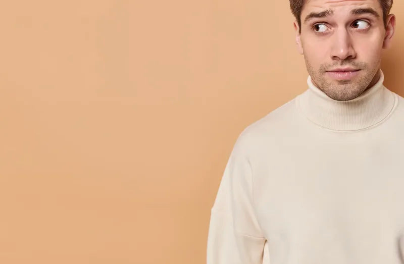 A man in a cream turtleneck looks to his right with a curious or uncertain expression, standing against a plain beige background. The left side of the image is empty, drawing focus to his face.