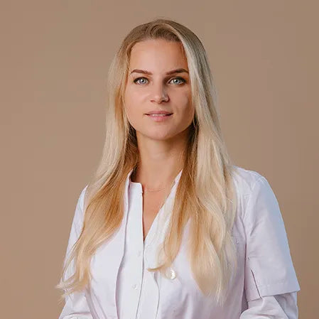 A woman with long blonde hair wearing a white buttoned lab coat stands against a plain beige background, looking forward with a neutral expression.