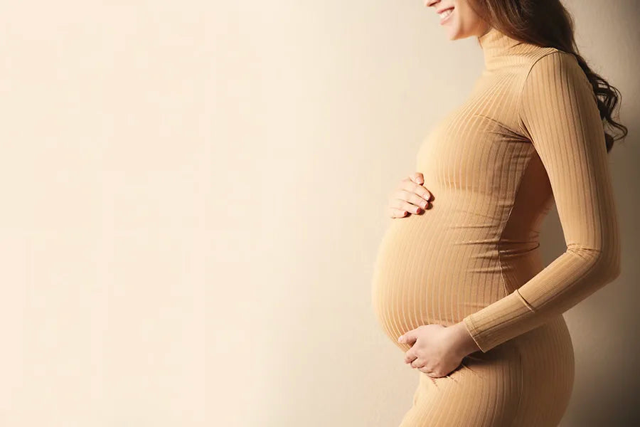A pregnant woman in a fitted, long-sleeve beige dress stands in profile, gently cradling her baby bump with both hands against a plain light background.