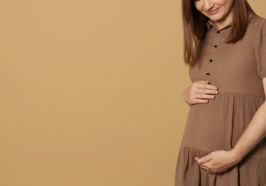 A woman in a brown dress stands against a beige background, gently cradling her baby bump and smiling softly. Only the upper half of her body is visible, with her face partially out of frame.