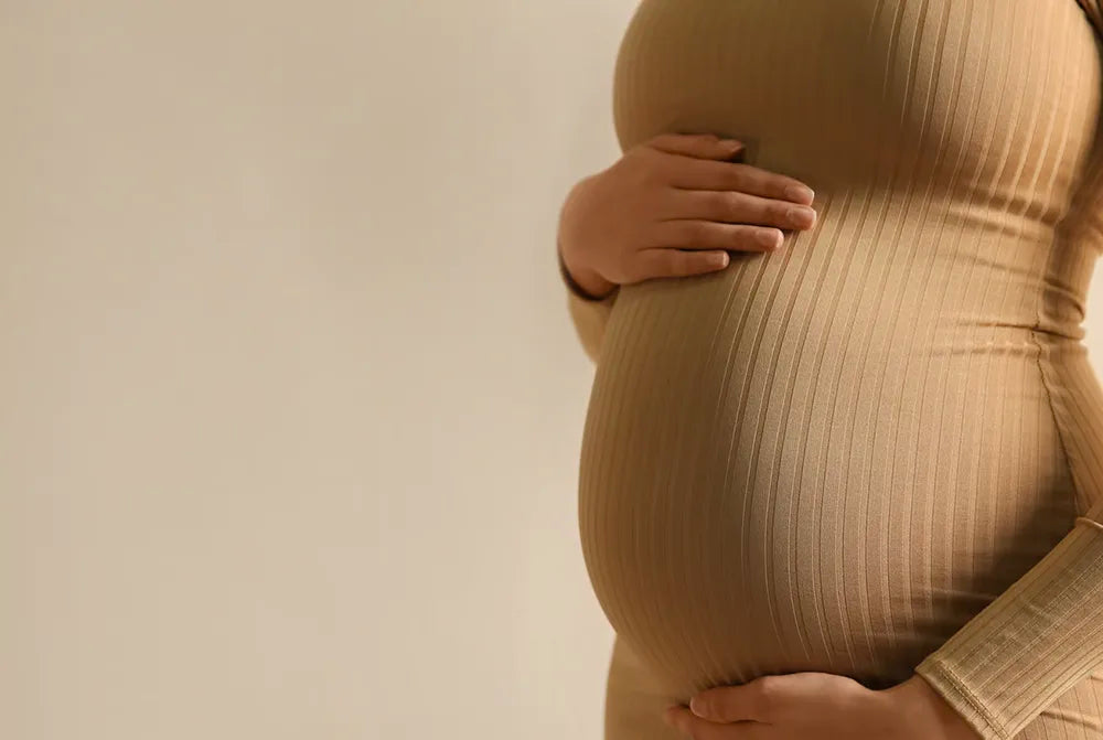 A person in a beige, ribbed dress holding their pregnant belly with both hands, standing against a plain, light-colored background.