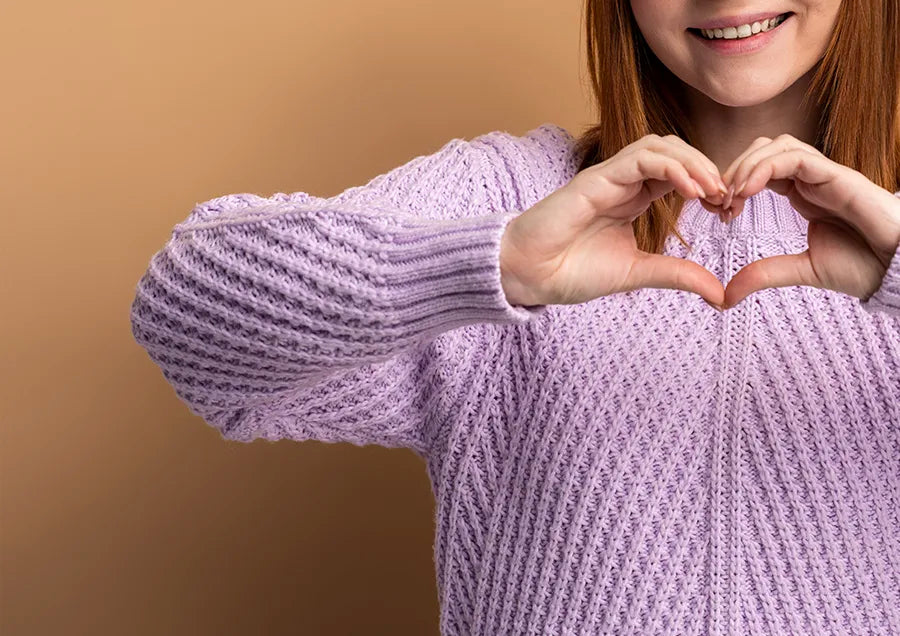 A person wearing a light purple sweater forms a heart shape with their hands. Only the lower half of their smiling face is visible, set against a plain beige background.