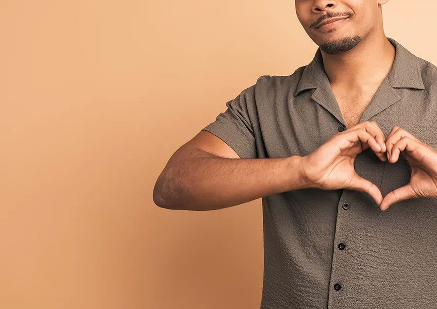 A person wearing a short-sleeved, button-up shirt forms a heart shape with their hands against a plain beige background. Only the upper body and one side of the face are visible.