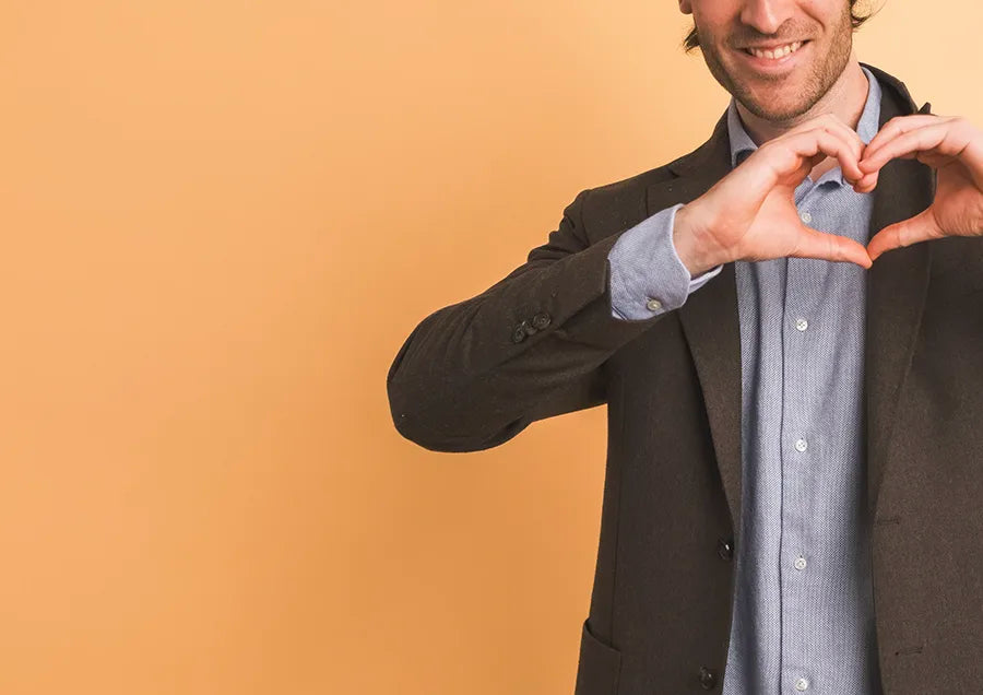 A man in a suit and blue shirt, smiling, forms a heart shape with his hands against a plain orange background. Only the upper half of his body is visible on the right side of the image.
