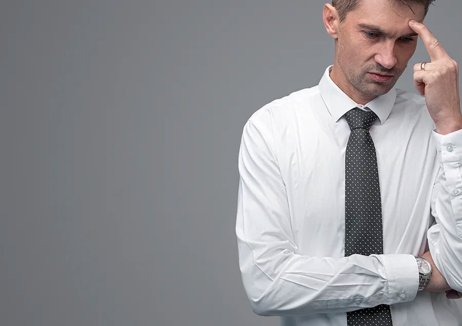 A man in a white dress shirt and black tie stands against a gray background, looking down thoughtfully with one hand touching his temple and the other arm crossed.