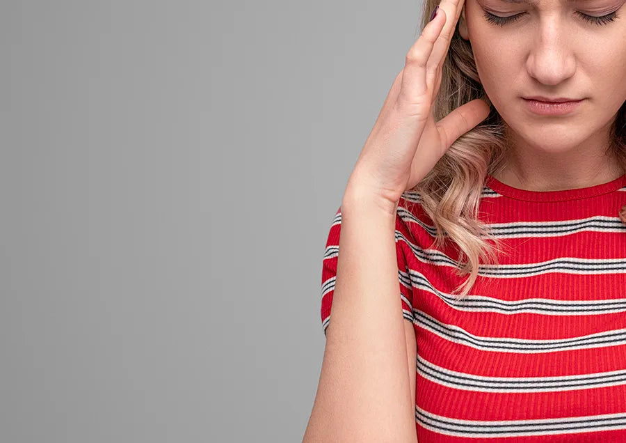A young woman wearing a red striped shirt touches her temple and closes her eyes, appearing to be in discomfort or pain, against a plain gray background.