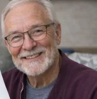 An older man with gray hair, a beard, and glasses smiles warmly at the camera. He is wearing a maroon sweater over a gray shirt and is holding a piece of paper. The background is softly blurred.
