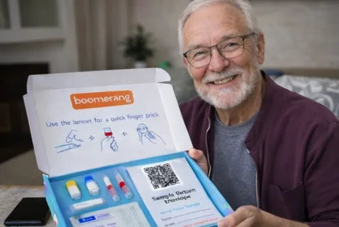 An older man with glasses and a beard smiles while holding an open home health test kit that includes vials, a lancet, instructions, and a sample return envelope.