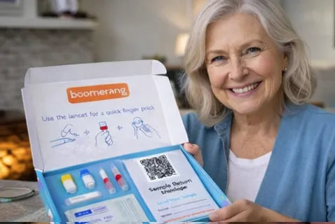 An older woman with gray hair smiles while holding an open home blood test kit labeled "boomerang," displaying medical supplies and instructions inside the box.