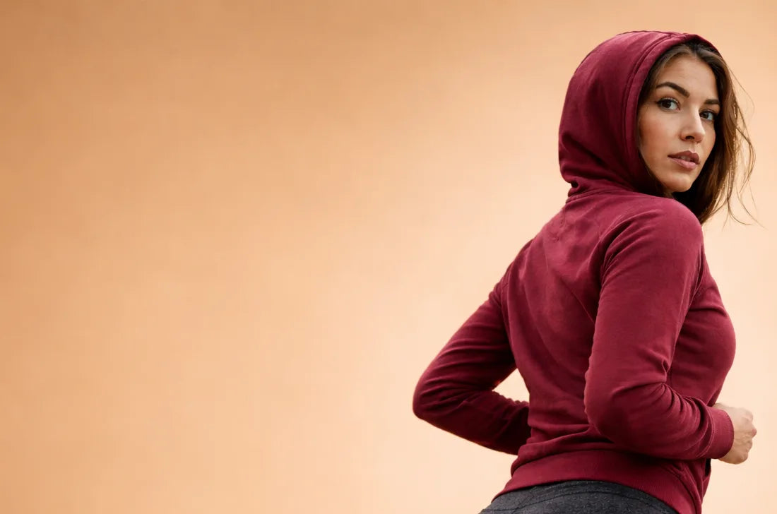 A woman in a maroon hoodie stands sideways, looking over her shoulder at the camera, against a plain beige background.