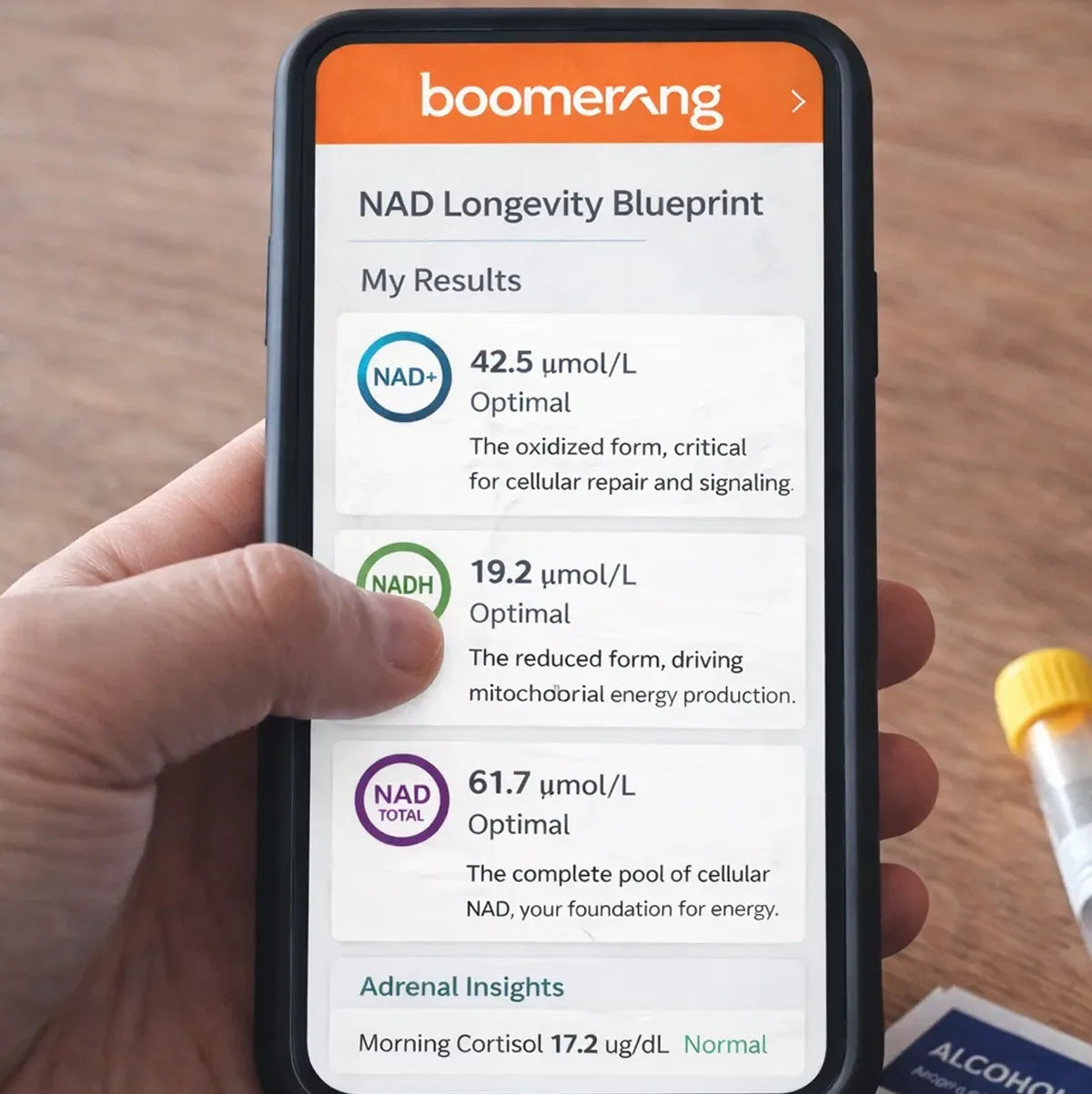 A hand holds a smartphone displaying lab results from the Boomerang app, showing optimal NAD+ (42.5 µmol/L), NADH (19.2 µmol/L), and NAD Total (61.7 µmol/L) levels, along with normal cortisol results.