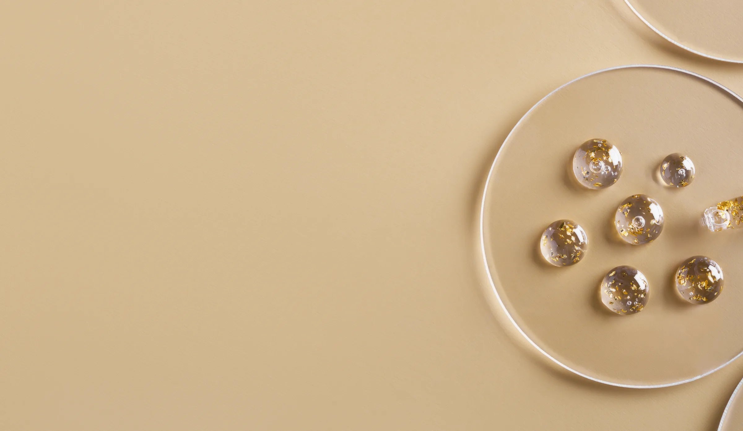 Clear round beads with gold flakes are arranged in a glass dish on a beige background, with a few beads outside the dish. The composition is minimal and elegant, emphasizing simplicity and light.