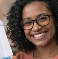 A woman with curly hair and glasses smiles while holding up a white and blue rectangular object.