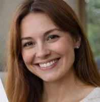 A young woman with long brown hair smiles warmly at the camera. She is indoors, and natural light highlights her friendly expression.