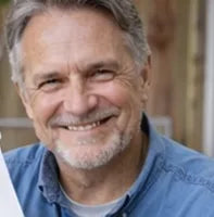 A smiling older man with gray hair and a beard, wearing a blue collared shirt, is pictured in an outdoor setting with a blurred wooden background.