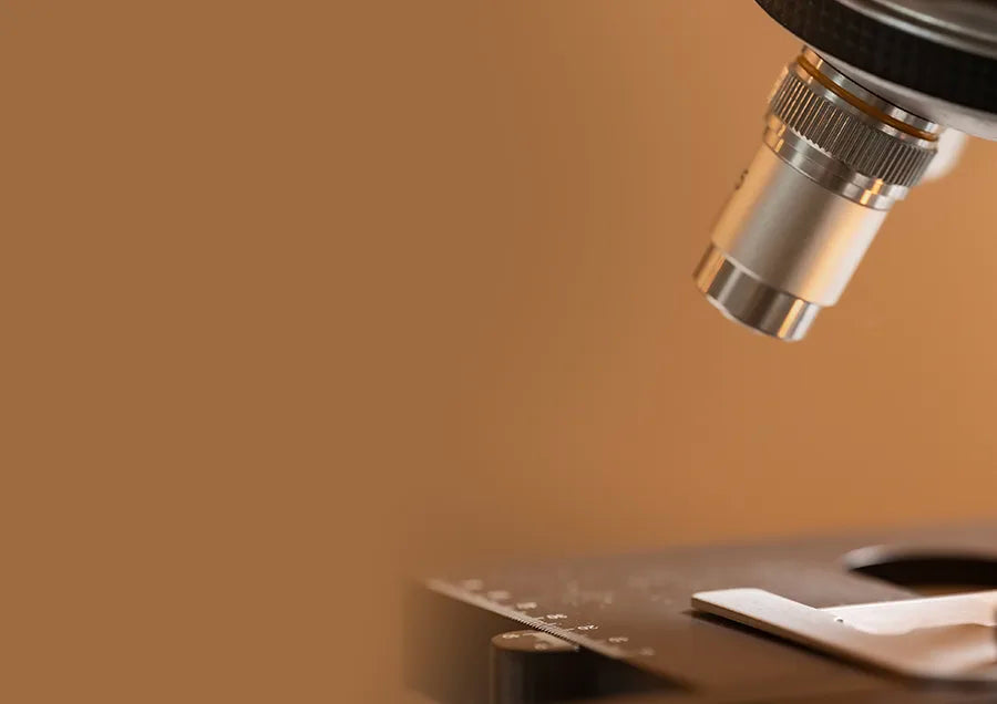 Close-up view of a microscope lens focused on a glass slide, with a warm brown background and empty space on the left side.