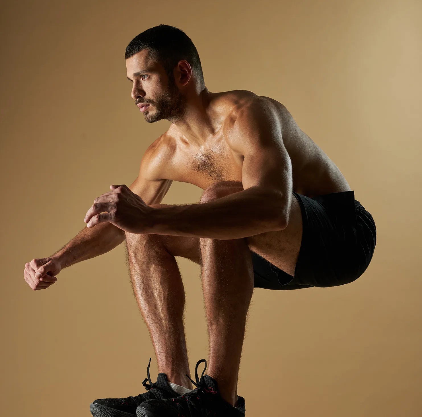 A shirtless man in black shorts and sneakers performs a squat jump against a plain tan background, with arms bent and knees pulled up toward his chest.