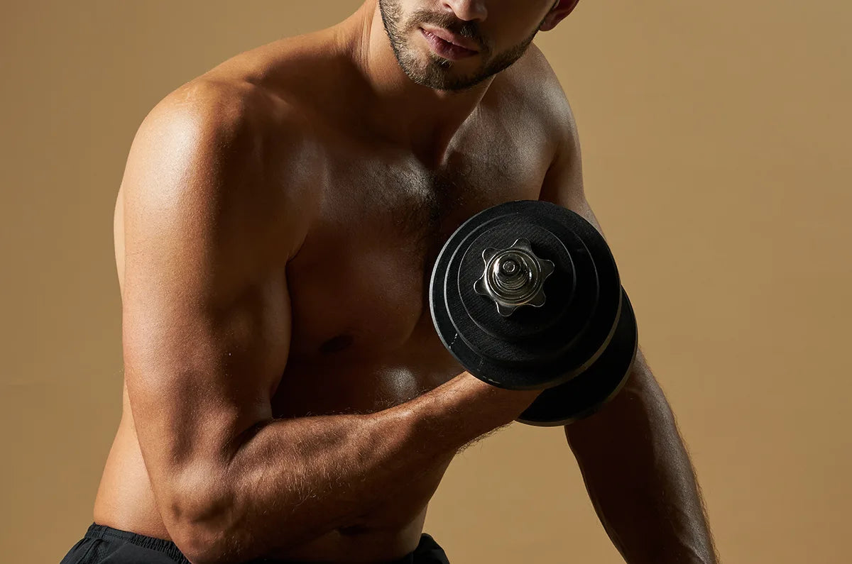 A shirtless man with a beard is lifting a black dumbbell with his right arm, showing defined muscles against a plain beige background.