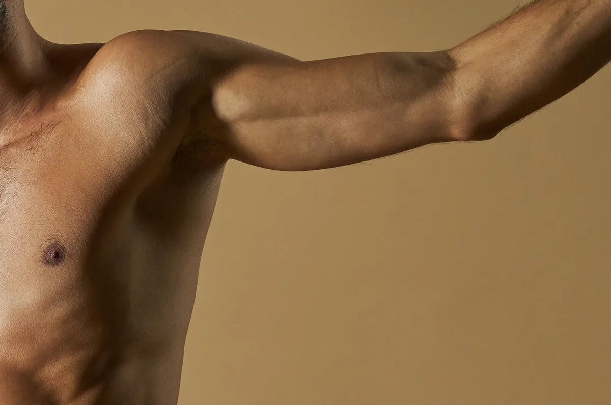 A close-up of a shirtless person’s upper torso and outstretched arm against a beige background. Only the left side of the chest and arm are visible.