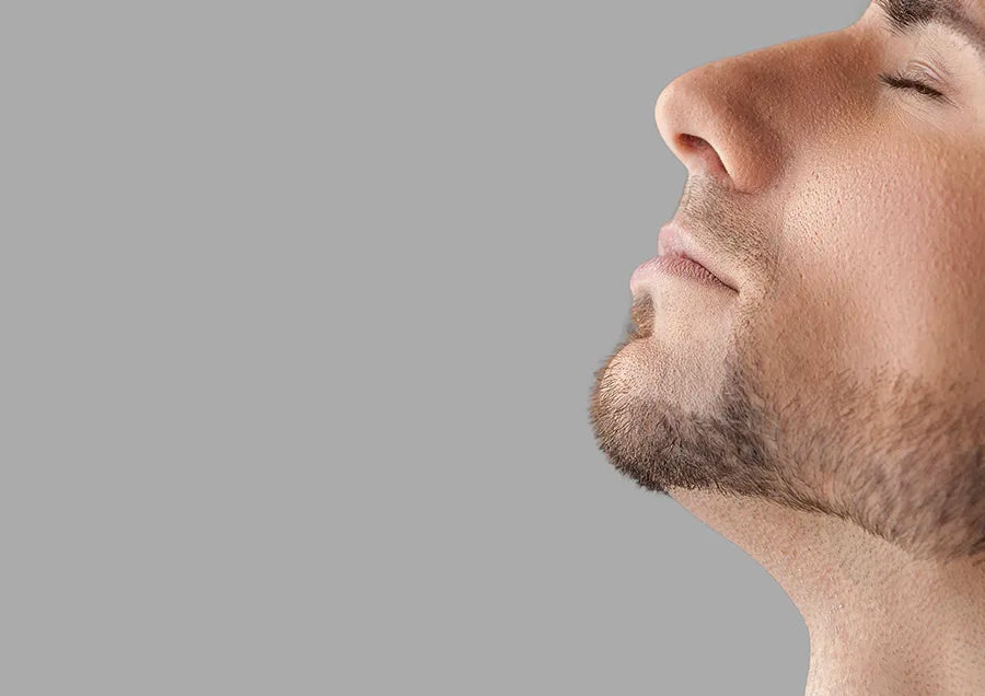 Close-up side profile of a young man's face with light facial hair, closed eyes, and relaxed expression, set against a plain gray background.