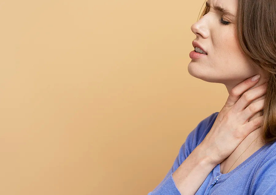 A woman in a blue top holds her throat and winces in pain, possibly experiencing discomfort or a sore throat, against a plain beige background.